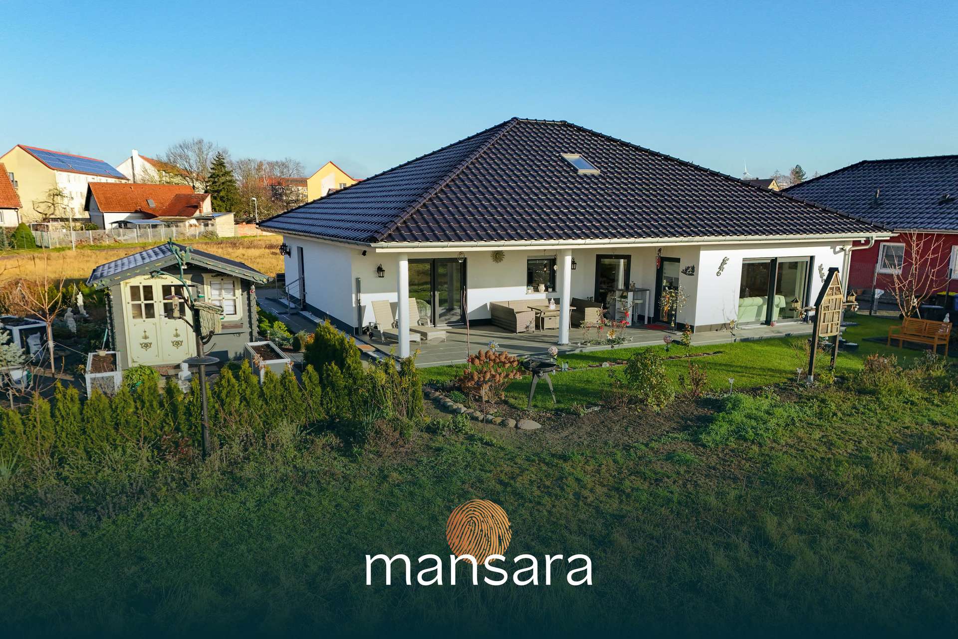 Ein modernes, einstöckiges Haus mit Terrasse, umgeben von Garten und einer kleinen Gartenlaube.