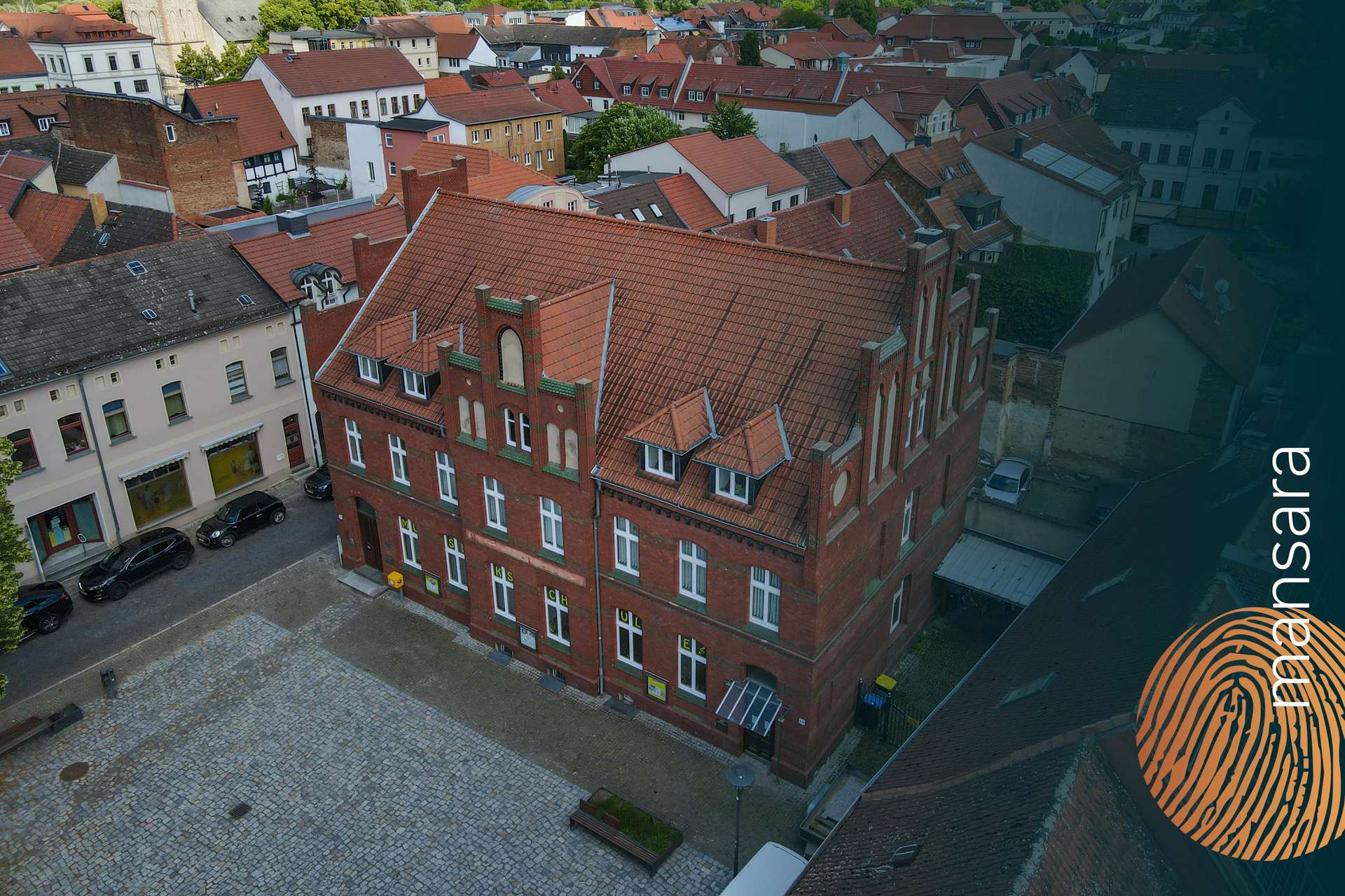 Luftaufnahme eines roten Backsteingebäudes mit Giebeldach, umgeben von städtischen Wohnhäusern und gepflastertem Platz.