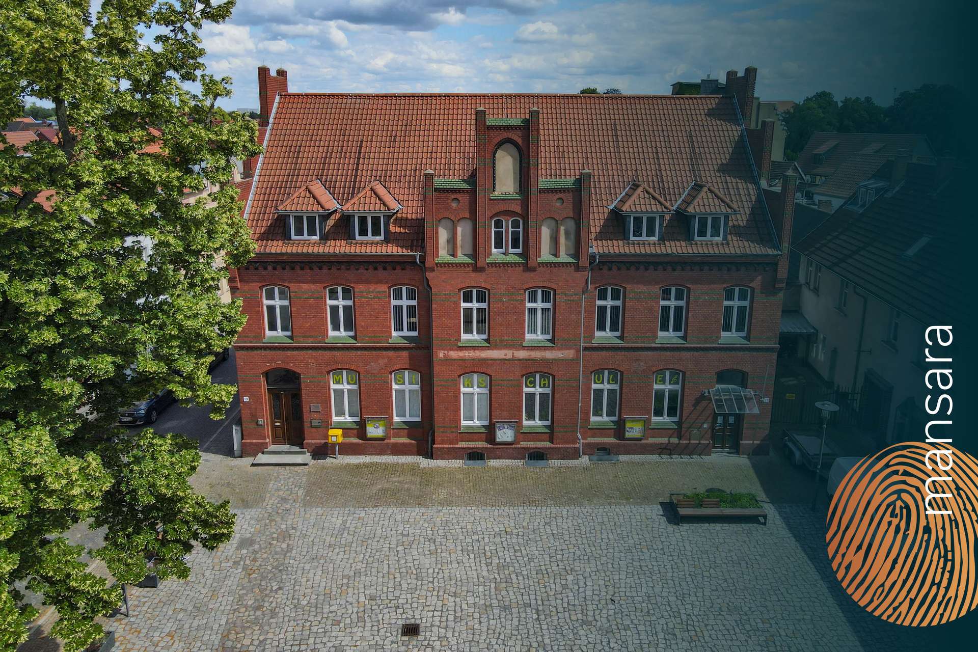 Historisches, rotes Backsteingebäude mit Giebeldach und großen Fenstern, umgeben von einer gepflasterten Fläche.