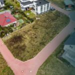 Ein unbebautes Grundstück in einer Wohngegend, umgeben von Häusern und Straßen, mit braunem Gras und vereinzelten Steinen.