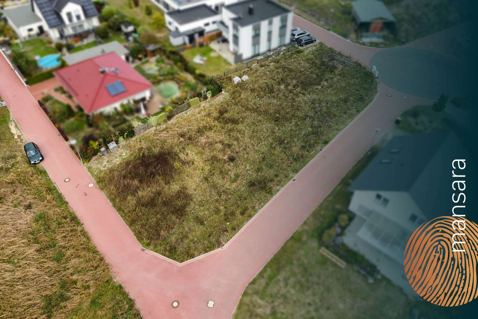 Ein unbebautes Grundstück in einer Wohngegend, umgeben von Häusern und Straßen, mit braunem Gras und vereinzelten Steinen.