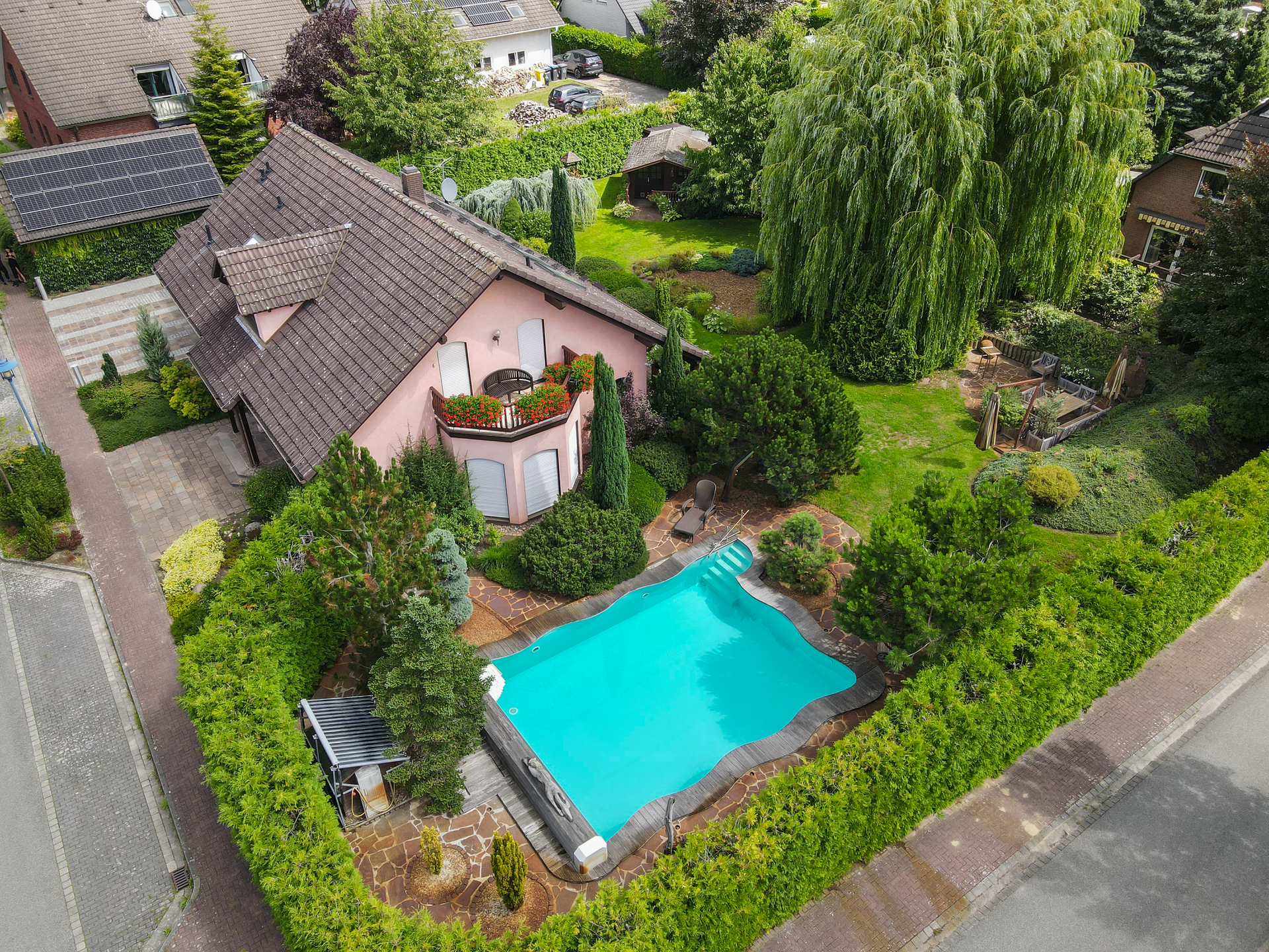 Ein Haus mit rotem Dach umgeben von Bäumen und einem blauen Pool im Garten, umgeben von gepflegten Hecken.