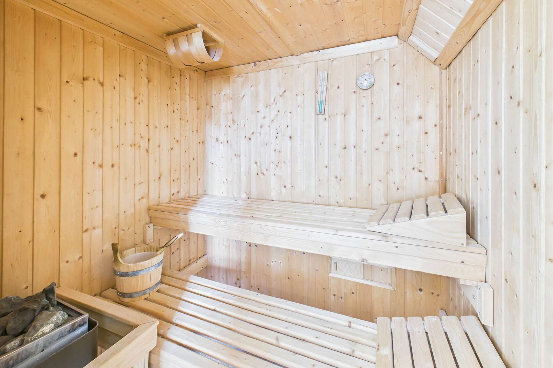 Innenansicht einer Sauna mit Holzverkleidung, Bänken, einem Eimer und einem Thermometer an der Wand.