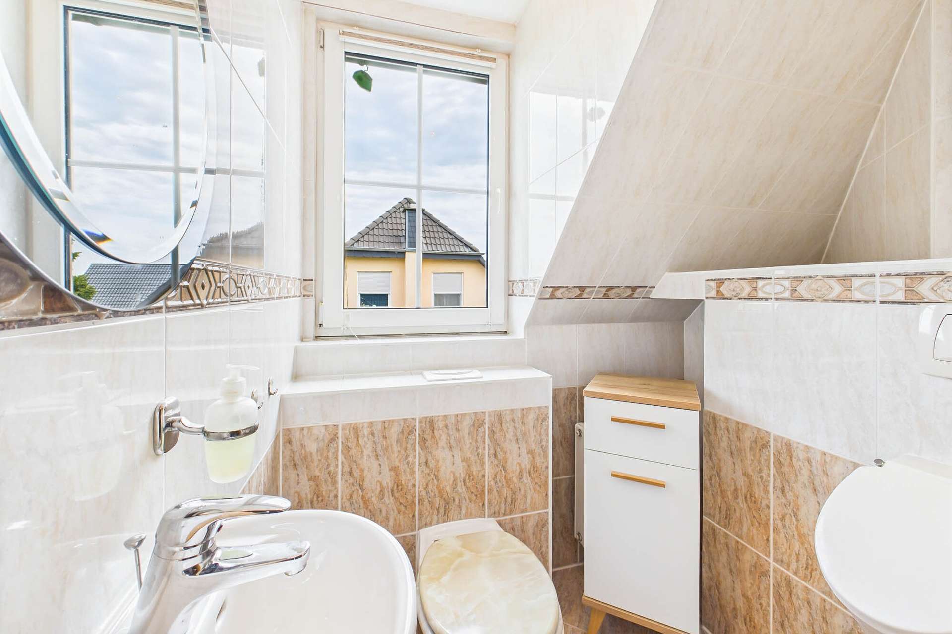 Badezimmer mit Fenster, Waschbecken, WC und Holzschrank, helle Fliesen und schräger Decke.
