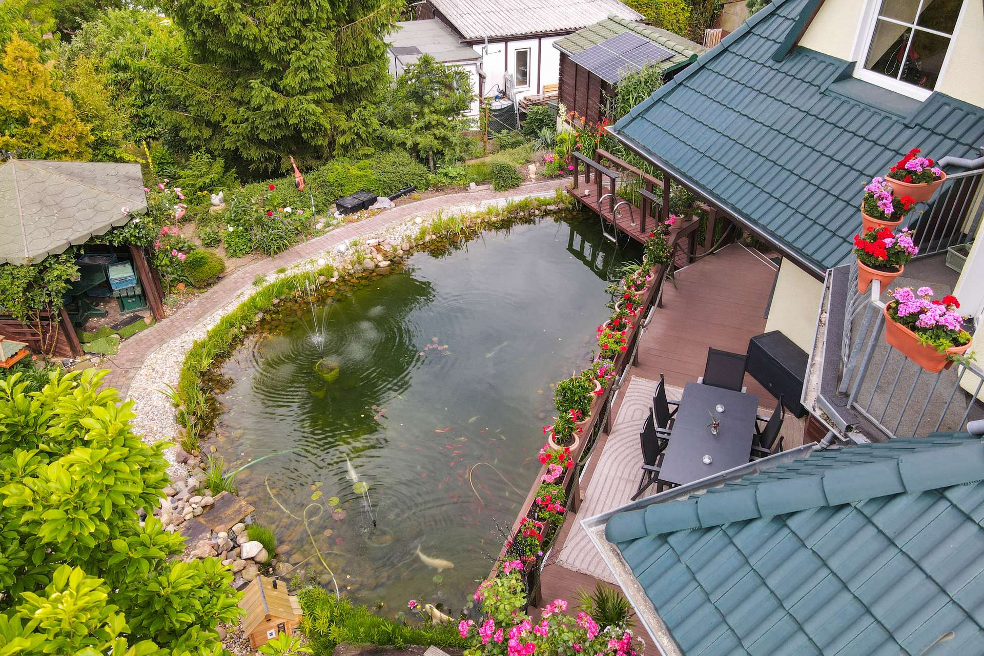 Ein Garten mit einem Teich, umgeben von Blumen und einem Holzdeck, neben einem Haus mit einem grünen Dach.