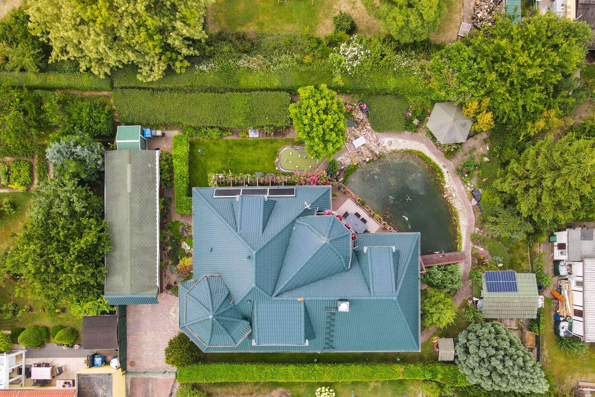 Luftaufnahme eines großen Hauses mit grünem Dach, umgeben von Gärten und einem Teich.