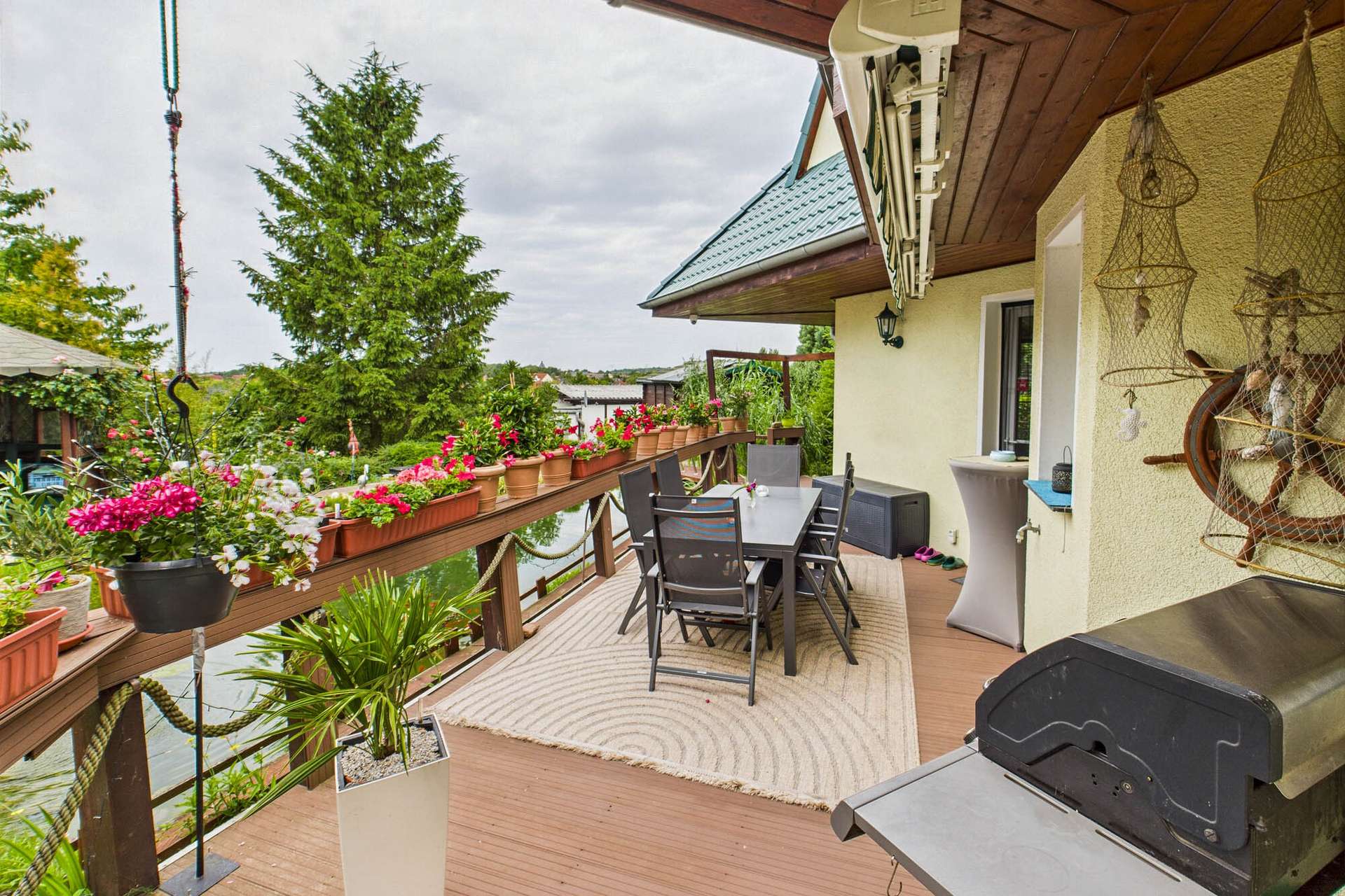 Terrasse mit Gartenmöbeln, Blumenkästen und Grill, umgeben von Pflanzen und Blick auf die Landschaft.