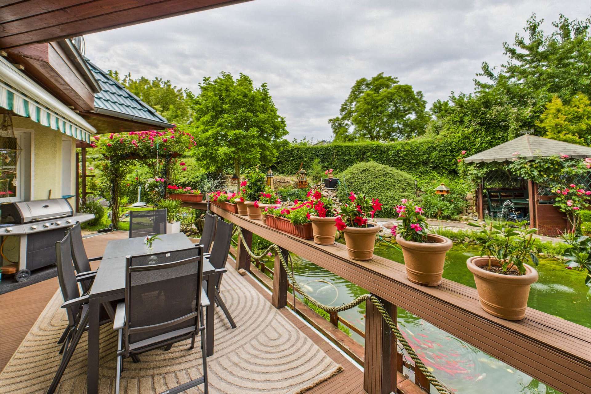 Terrasse mit Gartenmöbeln, blühenden Pflanzen und einem Teich im Hintergrund, umgeben von üppigem Grün.