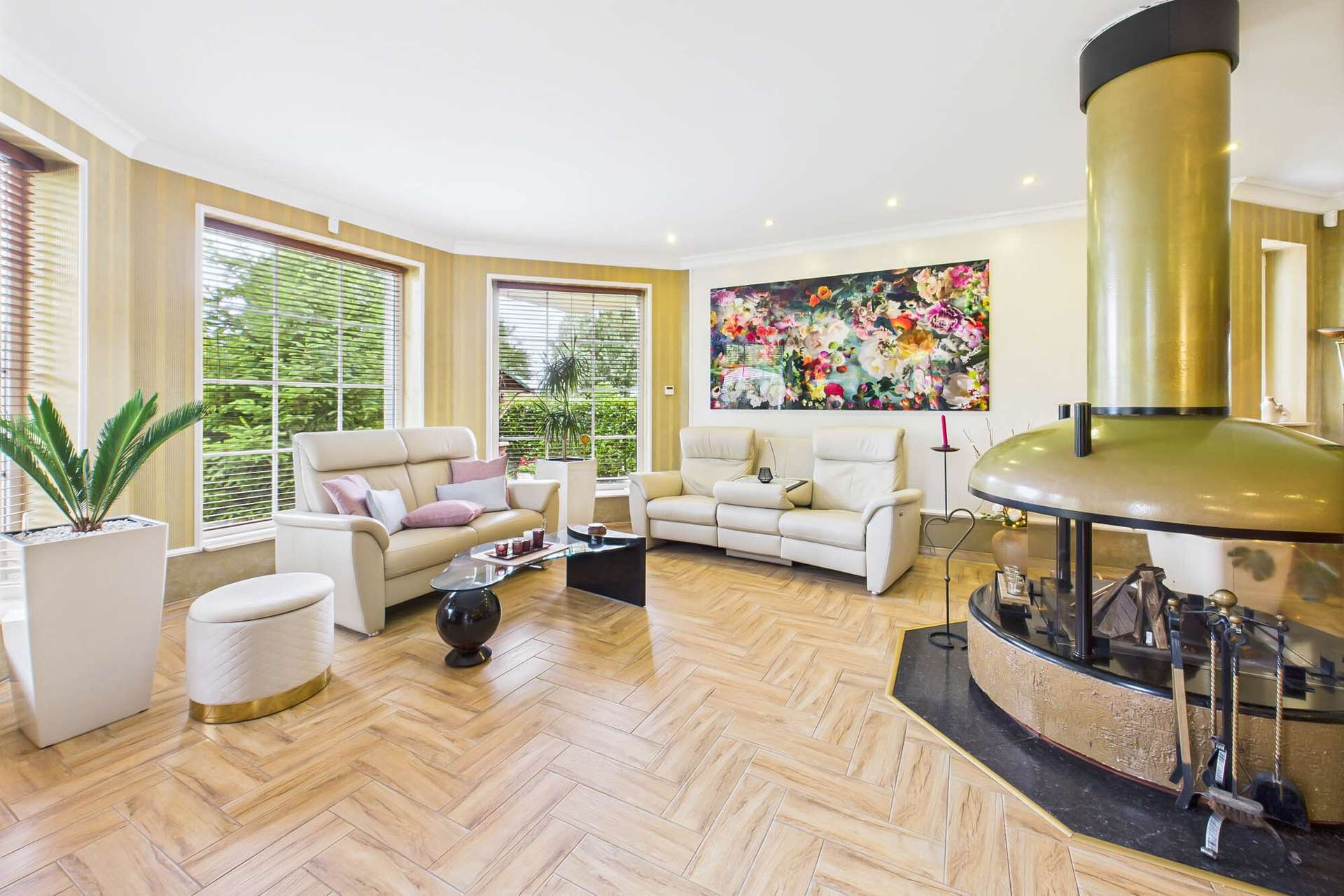 Heller Wohnraum mit zwei beigen Sofas, einem runden Tisch und einem goldenen Kamin, umgeben von großen Fenstern.