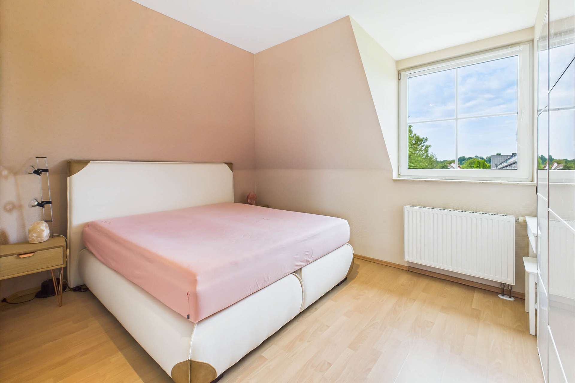 Schlafzimmer mit einem Bett, Holzfußboden und Fenster, das Tageslicht hereinlässt. Wände in sanften Farben.