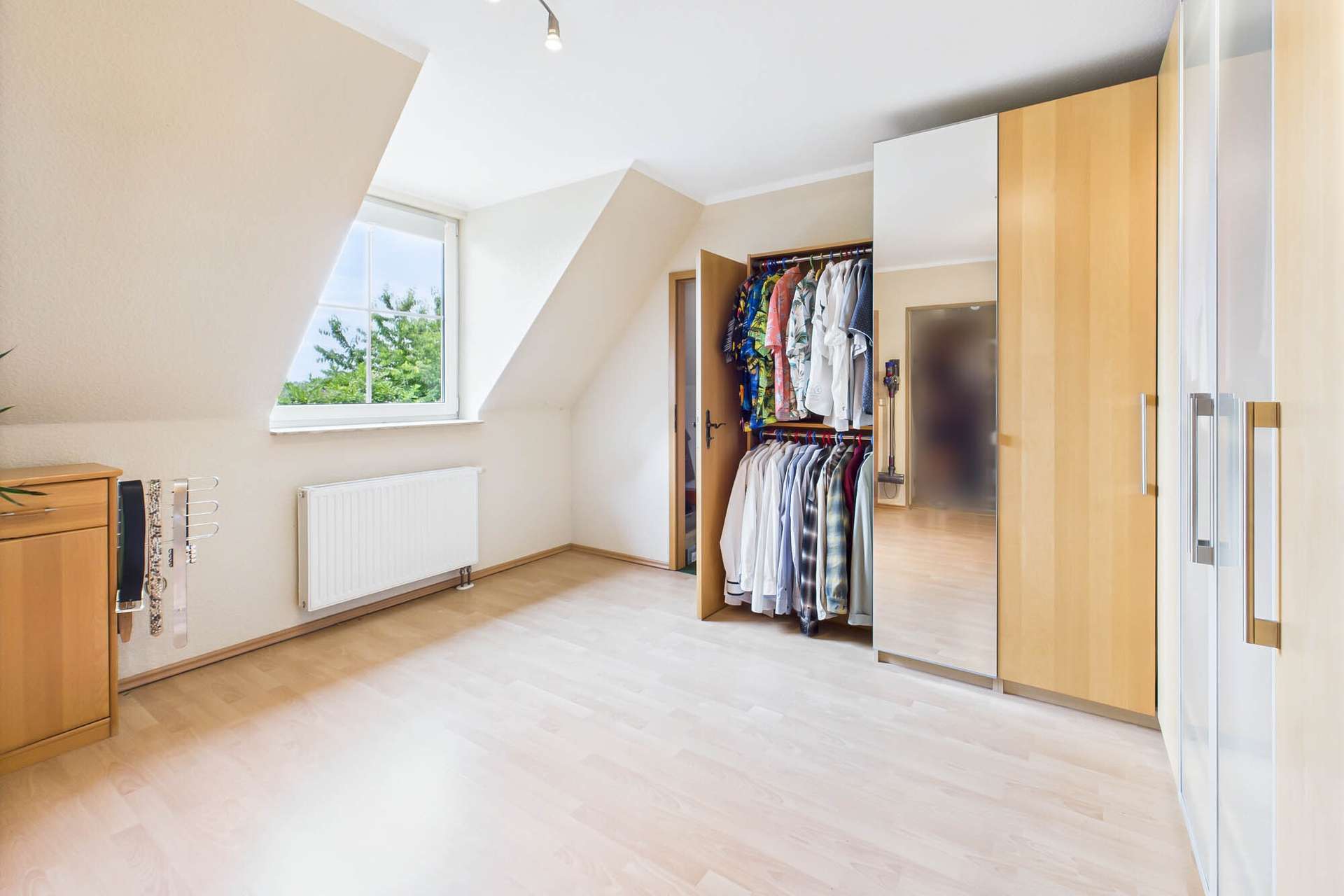 Helles Zimmer mit schräger Decke, Kleiderschrank, Spiegel und Fenster, das den Blick ins Freie bietet.