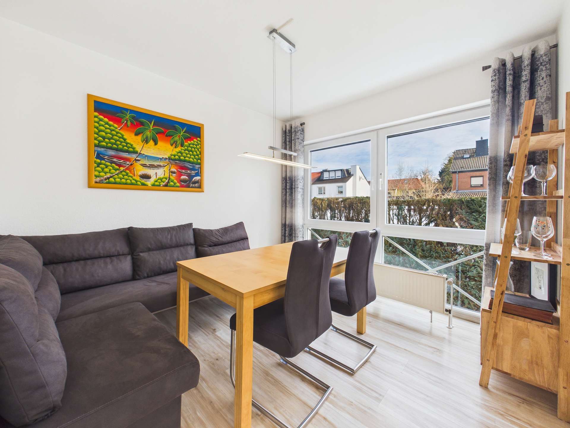 Helle Wohnküche mit Sofa, Esstisch und großen Fenstern, dekoriert mit einem bunten Gemälde.