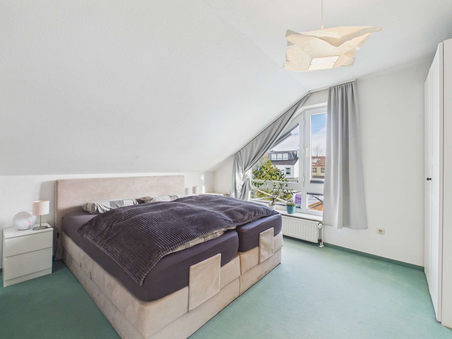Helles Schlafzimmer mit Doppelbett, grauer Bettdecke, Fenster mit Blick nach draußen und minimalistischer Einrichtung.