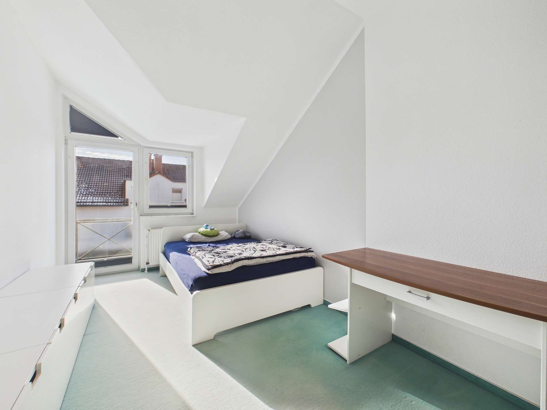 Helle Schlafzimmeransicht mit einem Bett, Schreibtisch und einem Fenster, das Tageslicht hereinlässt.