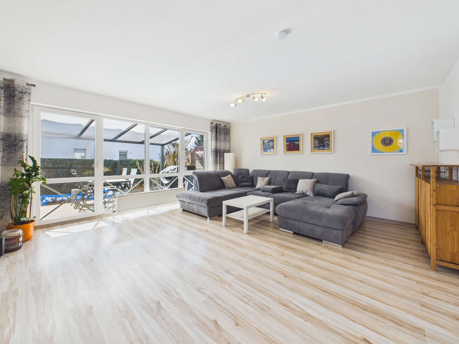 Heller Wohnraum mit grauem Sofa, weißem Tisch und großen Fenstern, die auf einen Garten blicken. Holzboden und Dekoration sichtbar.