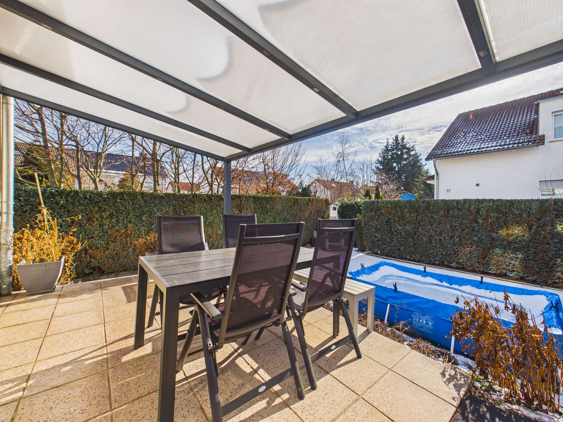 Terrasse mit Essgruppe aus schwarzen Stühlen und Tisch, umgeben von Hecken und mit einem blauen Planschbecken im Hintergrund.