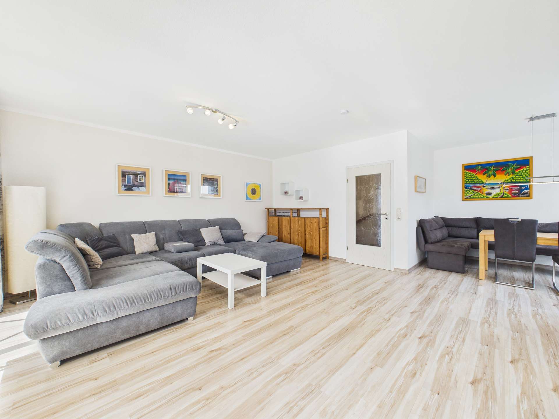 Heller Wohnraum mit grauem Sofa, weißem Tisch, Holzschränken und Esstisch, modernen Kunstwerken an den Wänden.
