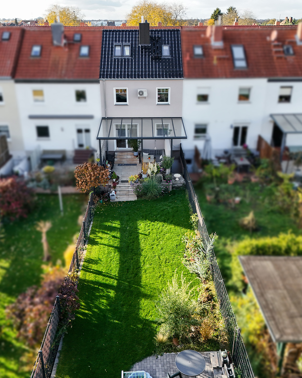 Hintergarten mit Rasen, Pflanzen und Terrasse, umgeben von WohnhÀusern. Sonnenlicht wirft Schatten auf den Boden.
