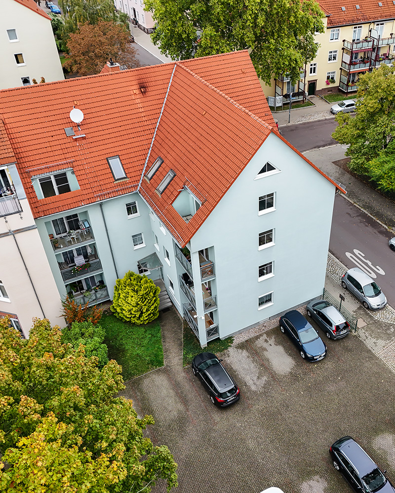Blick auf ein mehrstöckiges WohngebÀude mit rotem Dach, umgeben von BÀumen und parkenden Autos.