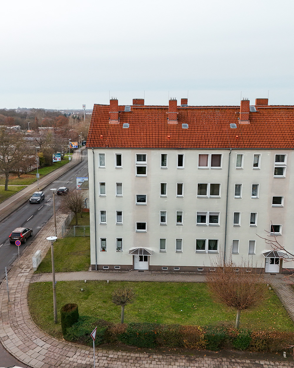 Mehrfamilienhaus mit rotem Ziegeldach, umgeben von einer GrĂŒnflĂ€che und StraĂenansicht im Hintergrund.
