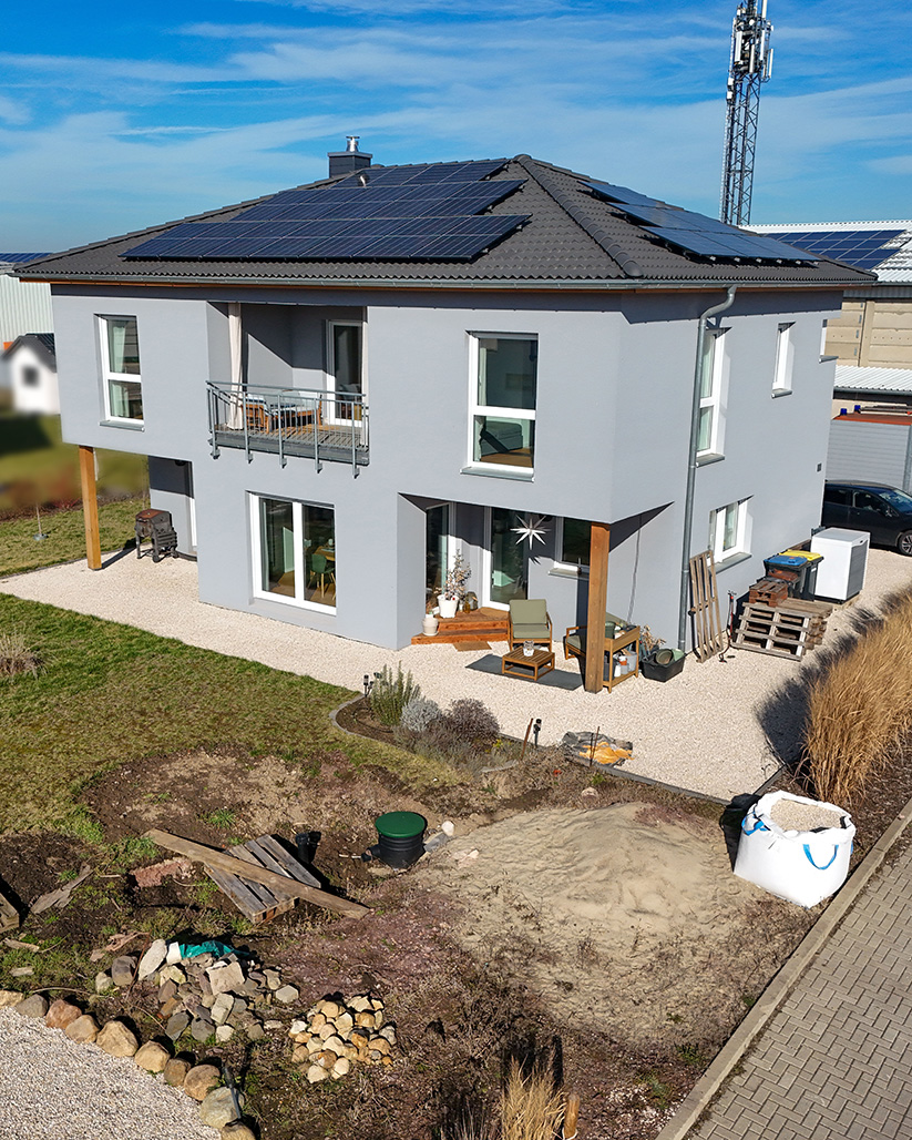 Zweistöckiges modernes Haus mit Solarpanelen, Garten und Terrasse in einer ländlichen Umgebung.