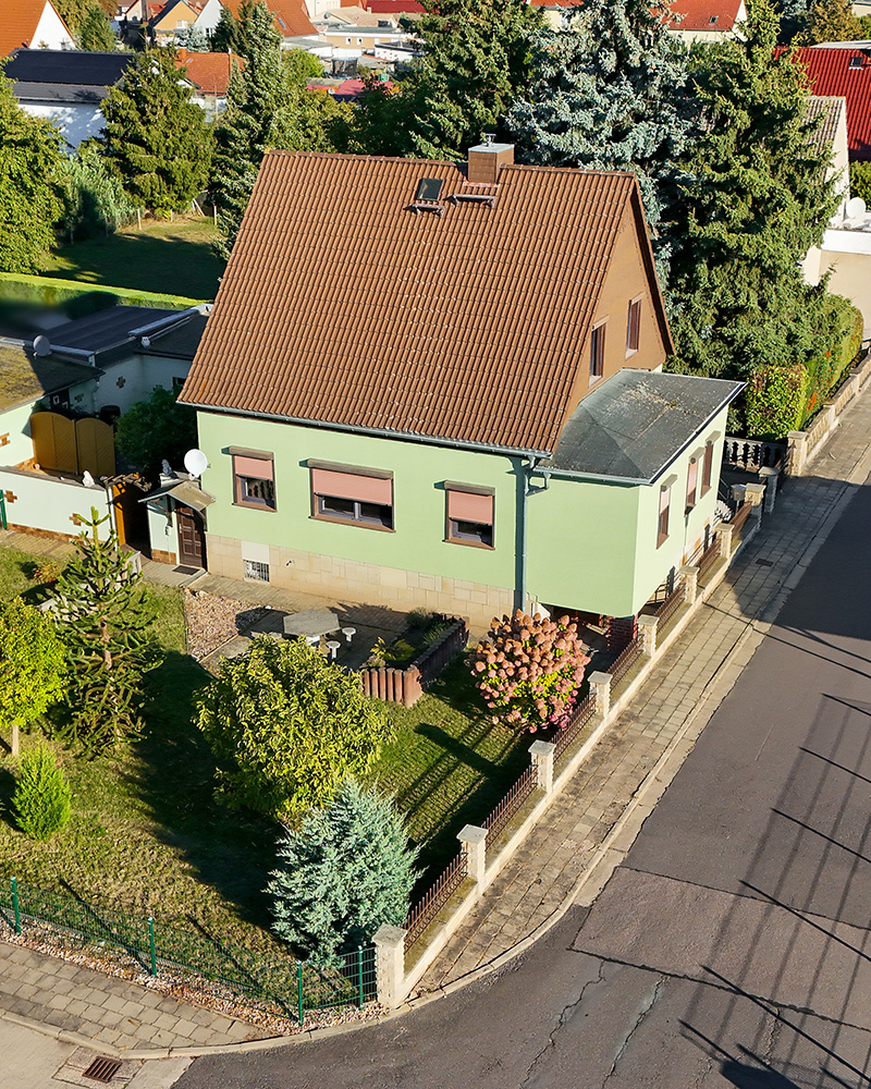 Ein grĂŒnes Einfamilienhaus mit einem roten Satteldach, umgeben von gepflegten GĂ€rten und BĂ€umen in einer ruhigen Nachbarschaft.
