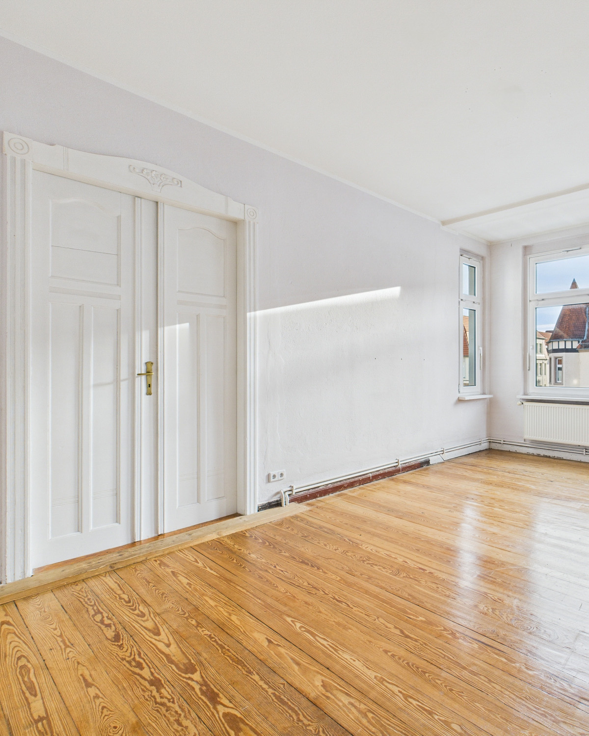 Heller Raum mit Holzboden, weiĂen WĂ€nden und doppelter ZimmertĂŒr, Fenster mit Blick nach drauĂen.