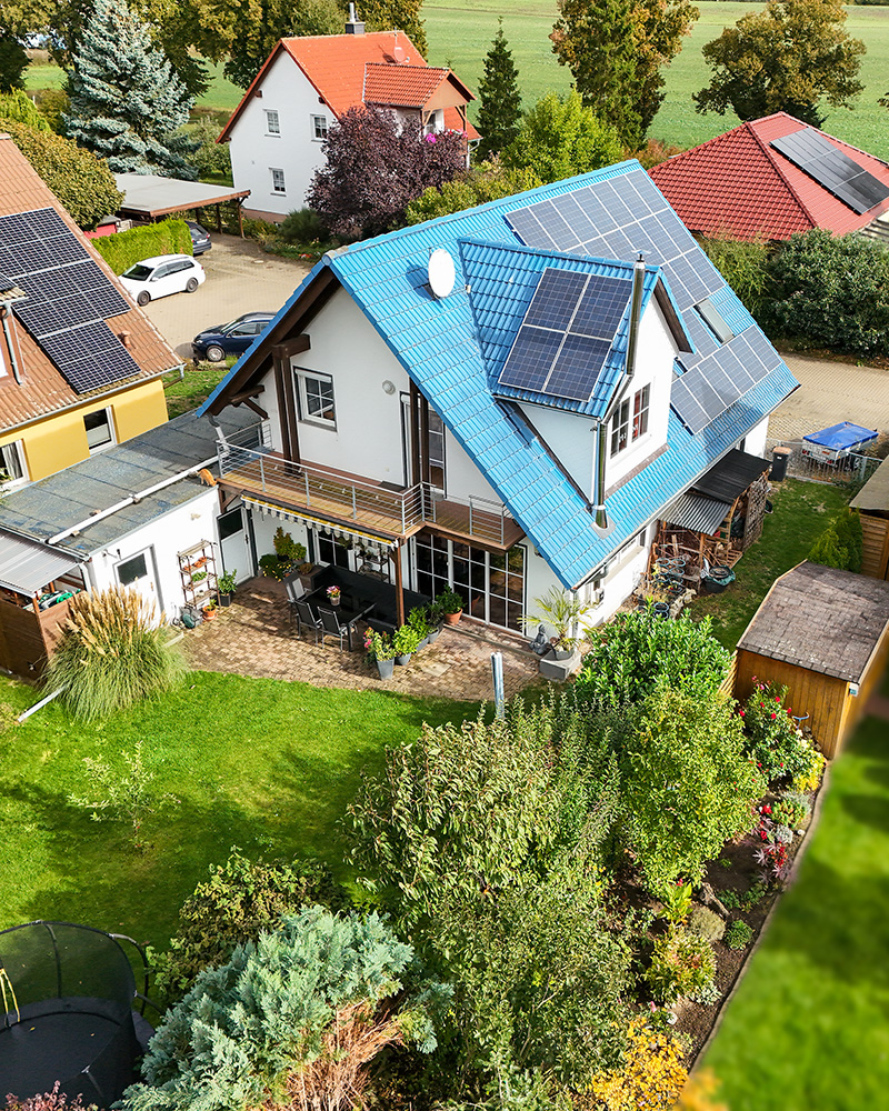Helles Haus mit blauer Dachziegeldeckung und Solarpanels, umgeben von einem gepflegten Garten und NachbarhÀusern.