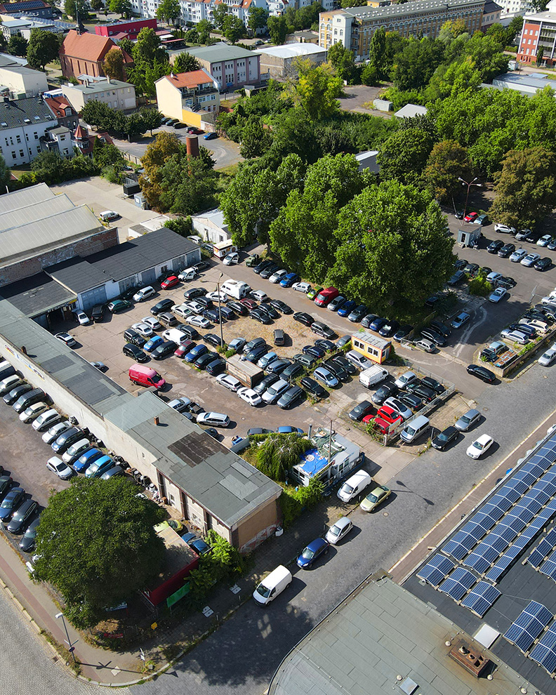 Luftaufnahme eines Parkplatzes mit zahlreichen Autos, umgeben von GebÀuden und BÀumen. Solarpanels im Vordergrund.