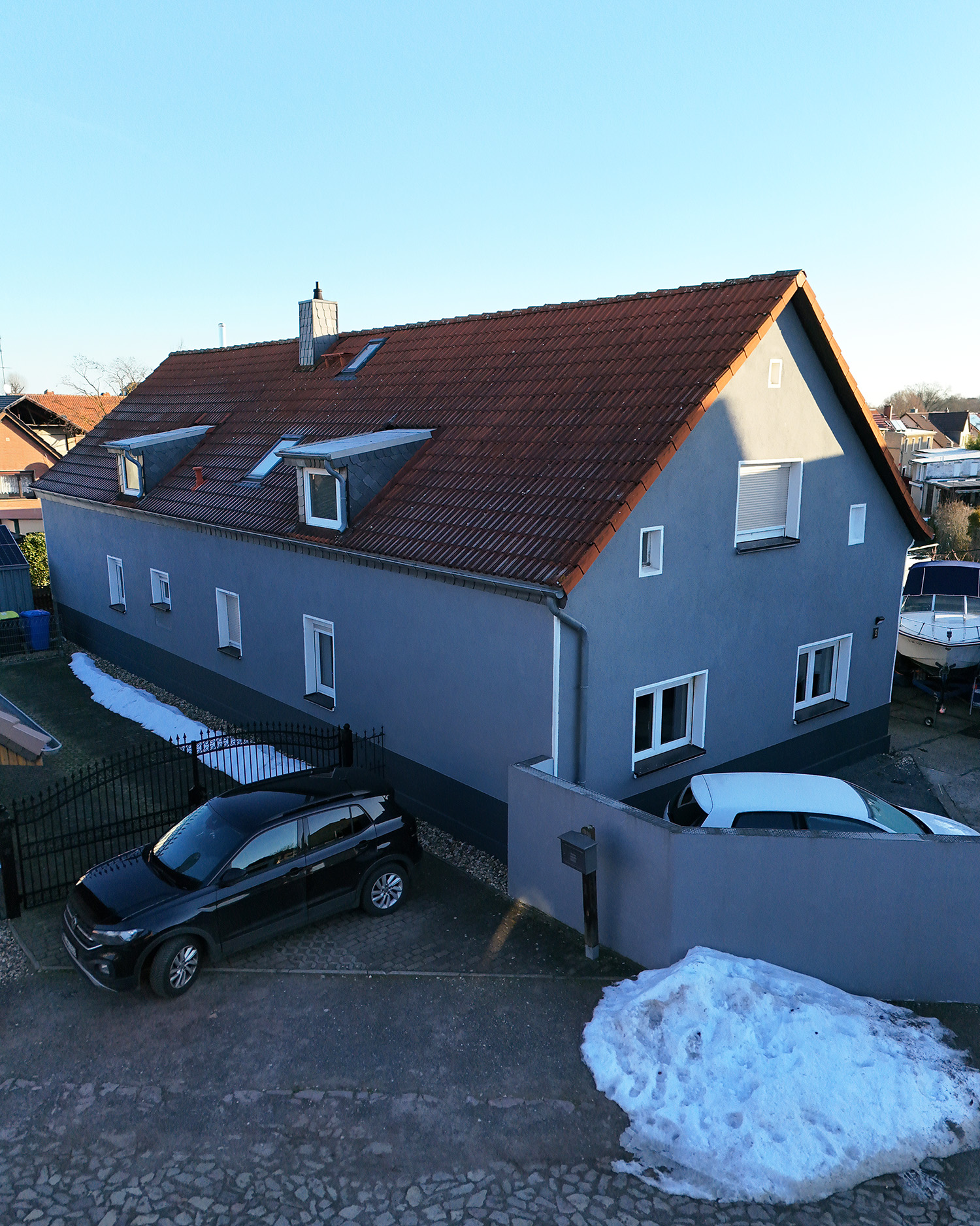 Ein graues Wohnhaus mit rotem Dach, umgeben von Schnee und zwei Autos auf einer Auffahrt.
