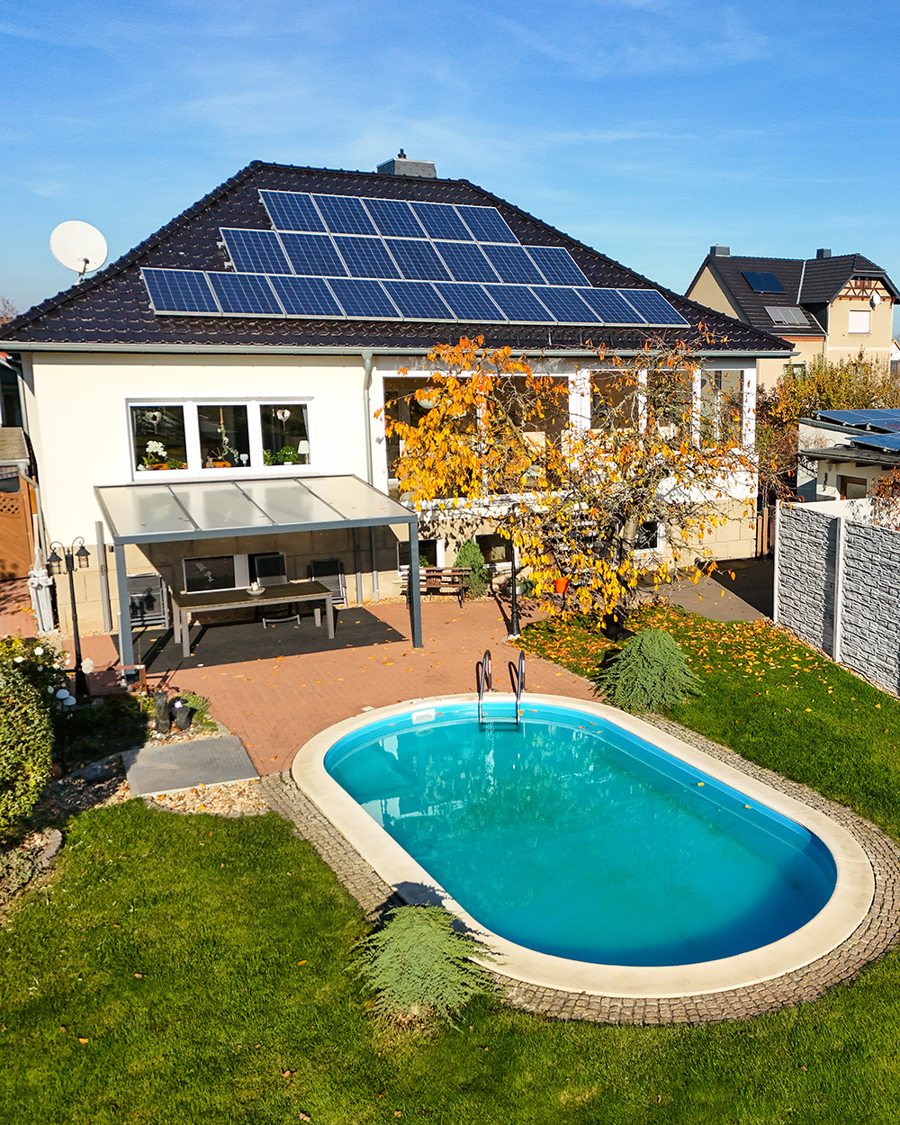 Modernes Haus mit Solarpanels, Garten und Pool, umgeben von herbstlichen BĂ€umen und klarem Himmel.