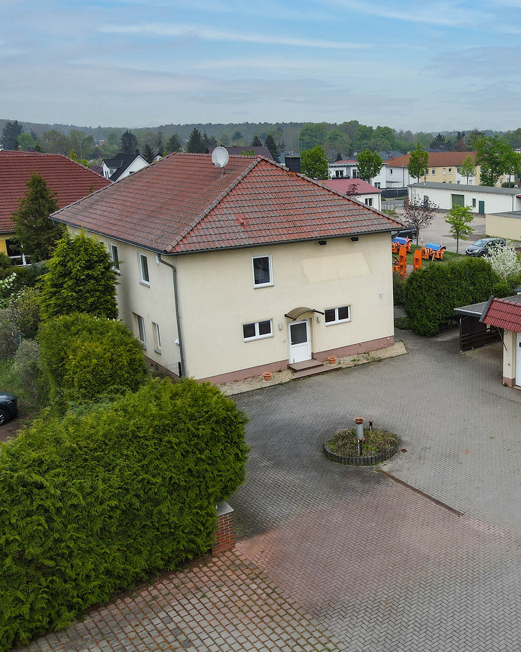 Ein zweigeschossiges, helles Wohnhaus mit rotem Dach, umgeben von GrĂŒnflĂ€chen und einer gepflasterten Einfahrt.