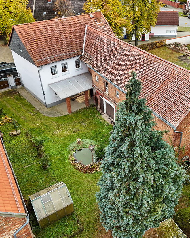 Luftaufnahme eines lĂ€ndlichen Anwesens mit zwei GebĂ€uden, Garten, Teich und groĂen Nadelbaum.