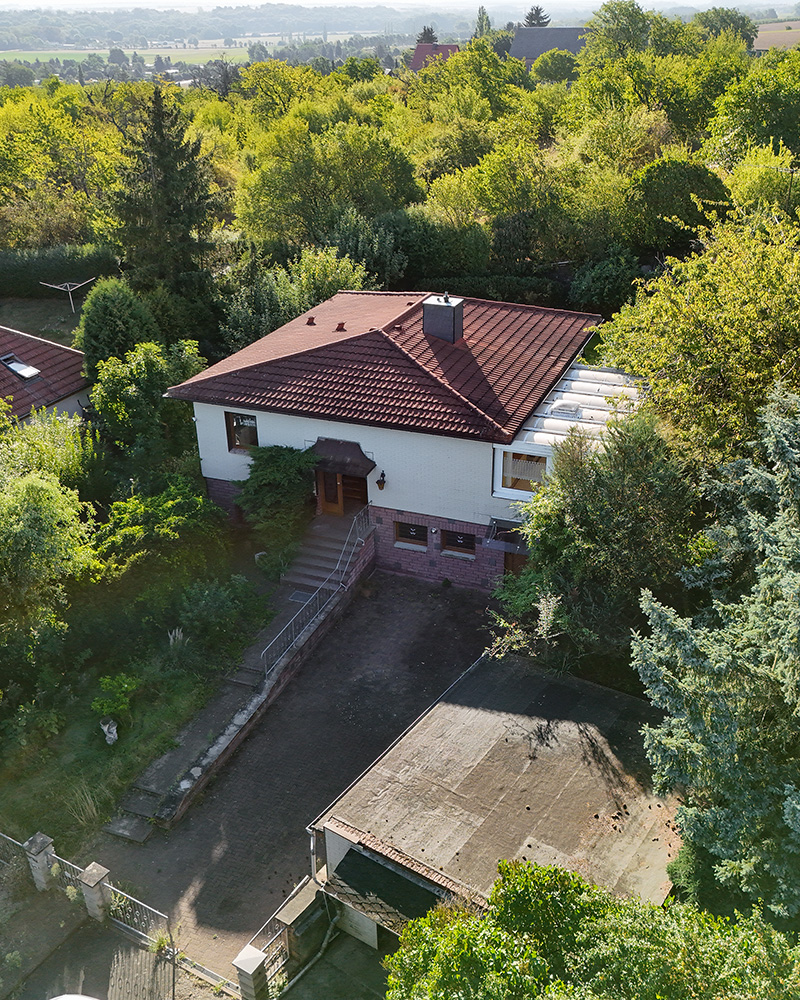 Ein zweigeschossiges Haus mit rotem Dach, umgeben von ĂŒppigem GrĂŒn und BĂ€umen, in einer ruhigen Umgebung.